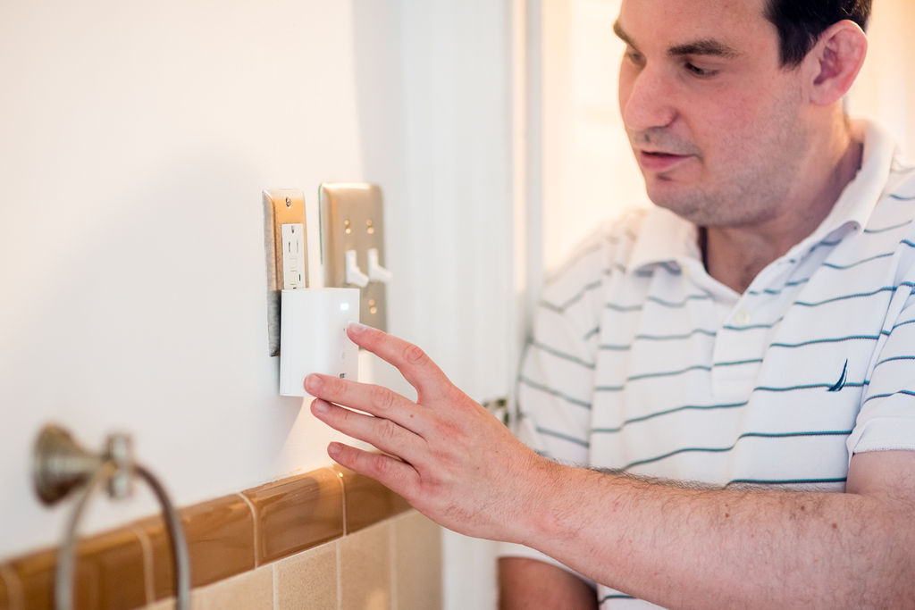 A white man stands in the doorway of a bathroom and touches a device plugged into the wall outlet. He is wearing a collared white shirt with thin blue stripes. The device is a small white box about three inches long with a small white light on the top front.