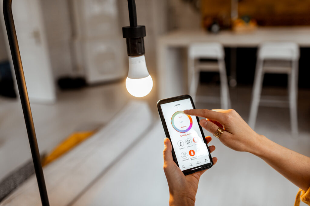 A white woman's hands hold a phone near a smart bulb. One hand taps the settings in the app.