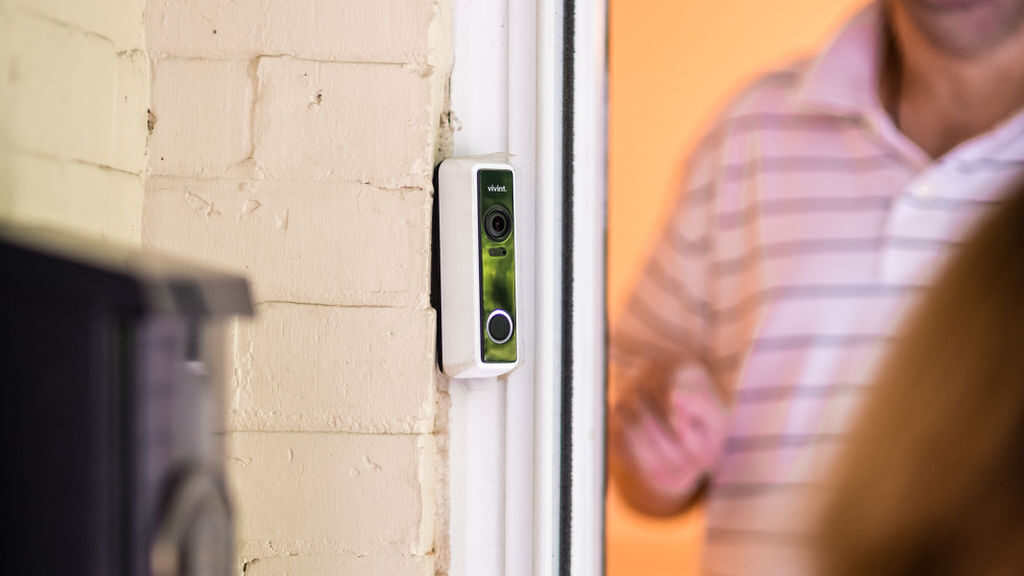 A smart doorbell on the frame of a door. A white man stands in the door frame and the back of a visitor's head is visible in the foreground.