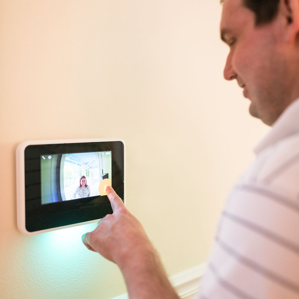 A white man taps a touch screen installed on a wall. The screen displays the view from his front doorbell, showing a woman smiling and standing outside.
