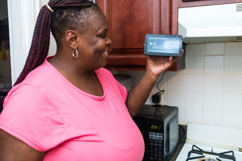 A black woman smiles and looks at a smart display she is holding up with one hand. She is wearing a pink short-sleeve dress and her hair is braided and pulled back. She is standing in a kitchen in front of a microwave and stovetop.
