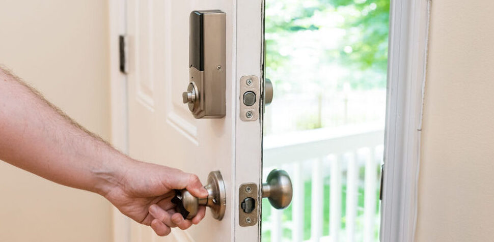 A front door is partially open to a bright front porch. A white man's hand rests on the door knob and there is a smart lock above it on the door.