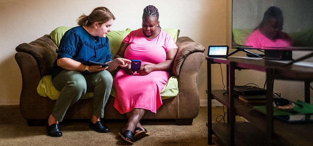 A white woman and a black woman sit on a couch together. The black woman holds a tablet and the white woman points to something on the screen.