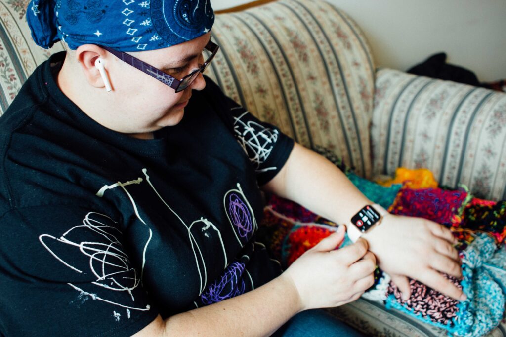 A white woman sits on a couch wearing a smart watch and cordless earbuds. She looks down and touches her watch with her other hand. She is wearing a blue bandana on her head, glasses, and a black t-shirt with hand-drawn white designs.