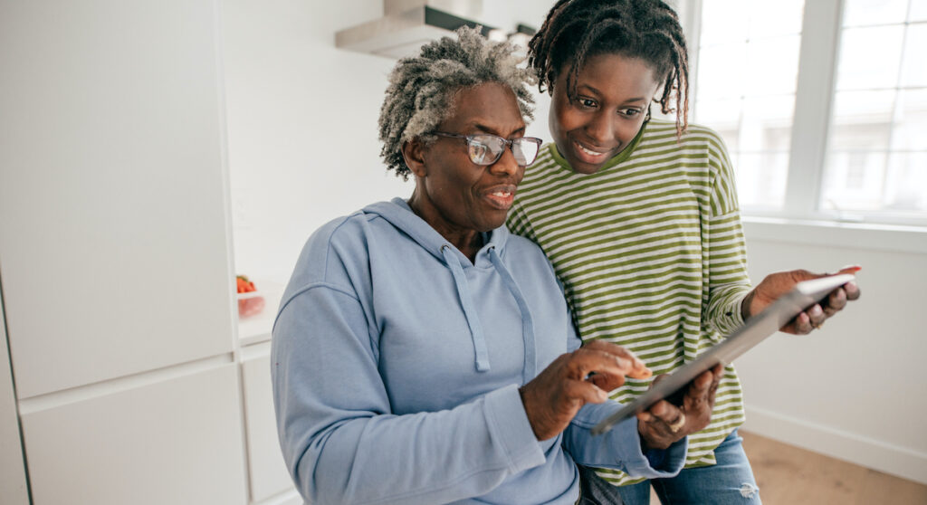 An older black woman and her young adult granddaughter smile while leaning over a tablet.